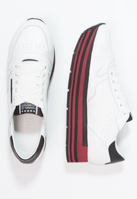 Scarpe sportive bianche con una parte superiore in pelle liscia, allacciatura tradizionale e una suola spessa a righe nera e bordeaux per un'altezza aggiuntiva.
