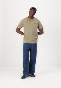 Maglietta verde oliva con logo nero, jeans in denim blu e sneakers blu con strisce bianche. Il modello indossa un cappellino blu navy. Vestibilità e texture casual.