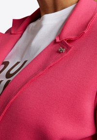 Klart rosa blazer i en texturerad tygkvalitet, prydd med en silverfärgad blompins. Den vita t-shirten under visar brun text i fet stil.