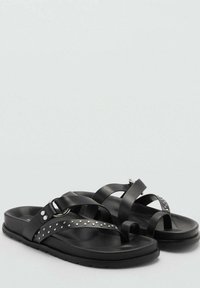 Černé ploché sandály s prstovými smyčkami, křižujícími se popruhy, stříbrnými hroty a stříbrným kováním na jednobarevném světle šedém pozadí.