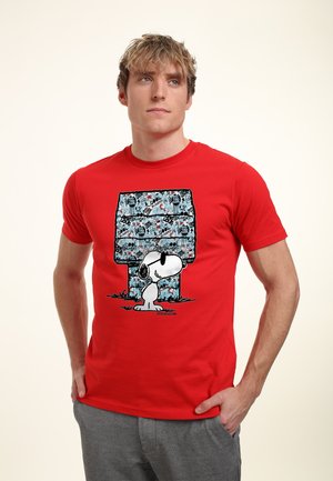 Giovane con una maglietta rossa con Snoopy e una cuccia blu coperta da testo in stile graffiti, in piedi con le mani nelle tasche.