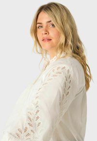 Blusa bianca con dettagli in pizzo lungo le maniche, dotata di un collo alto e una vestibilità ampia. Tessuto leggero con un aspetto testurizzato.