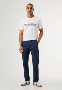 Hvit t-skjorte med svart og rødt trykk, kombinert med marineblå cargobukser med sidelommer. Fottøy inkluderer hvite og brune joggesko.