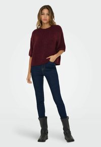 Una giovane donna sta in piedi con una mano in tasca, indossando un morbido pullover bordeaux, jeans skinny scuri e stivali neri fino al polpaccio, su uno sfondo bianco.