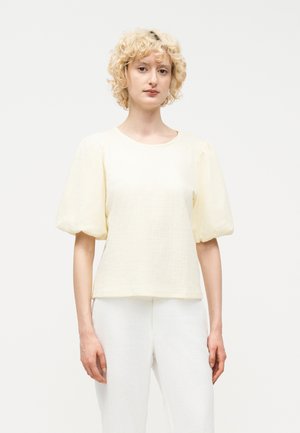 Blondynka o kręconych włosach, ubrana w kremową, teksturowaną bluzkę z bufkami na krótkich rękawach oraz białe spodnie, stojąca na tle białego tła.