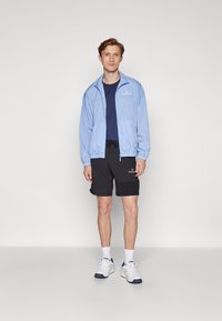 Lysblå zip-jakke, marineblå t-shirt, sorte shorts, hvide sneakers med marineblå detaljer. Casual atletisk tøj med elastisk talje og logoer.