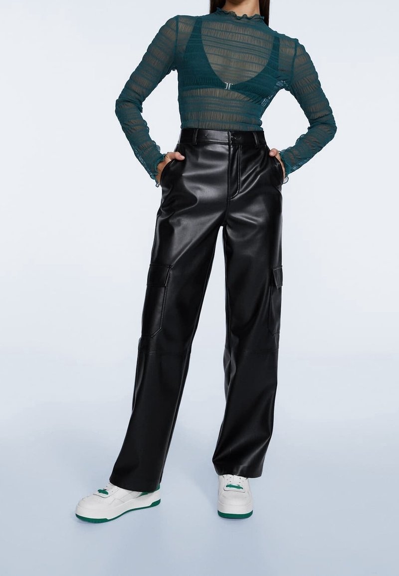 Femme portant un haut transparent à manches longues couleur sarcelle, un pantalon en cuir noir taille haute et des baskets blanches avec des semelles vertes.