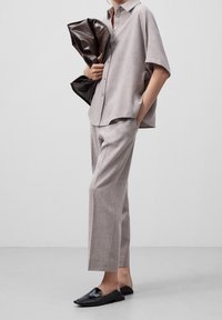 Lichtgrijze hemd met korte mouwen en knopen, gecombineerd met bijpassende broek, en zwarte loafers met strikdetail, terwijl een donkerbruine pouch tas wordt vastgehouden.