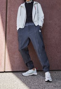 Lehká bílá bunda na zip s černými detaily, navlečená přes černé tričko. Temně šedé jogger kalhoty s elastickým pasem a bílým logem. Bílé tenisky.