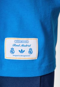 Blue cotton t-shirt with a white rectangular label featuring "Real Madrid," Adidas logo, and text "Designed in Herzogenaurach" in blue.