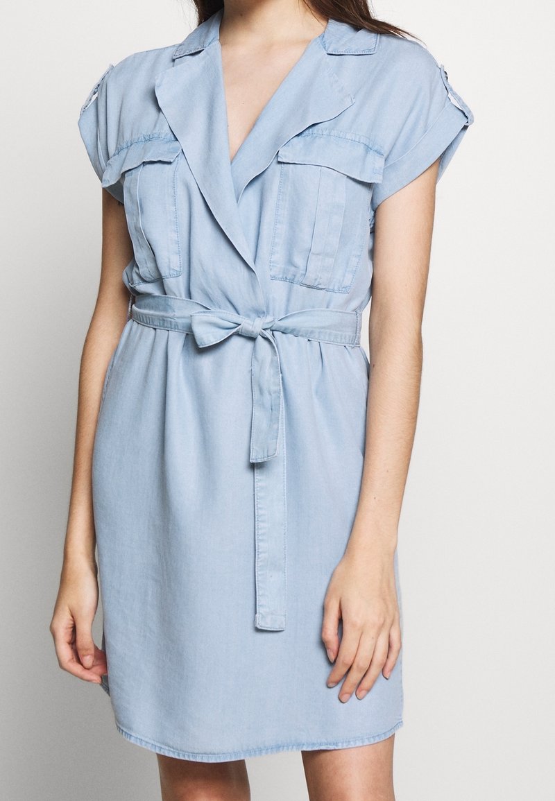 Robe chemise en denim bleu clair, manches courtes, deux poches poitrine, taille ceinturée et col cranté. Texture lisse avec un léger brillant.