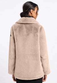 Beige nepbont jas met een gelapt kraag, zachte textuur en rechte snit. Heeft een patch op de linkerschouder en volledige lange mouwen.