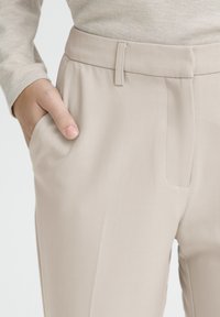 Un pantalon beige ajusté présente une texture lisse, des poches latérales et un devant plat, avec un léger éclat soulignant la qualité du tissu.