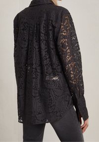 Camicia nera in pizzo con maniche lunghe, motivo floreale intricato, apertura frontale con bottoni e orlo curvato. Texture trasparente e vestibilità rilassata.