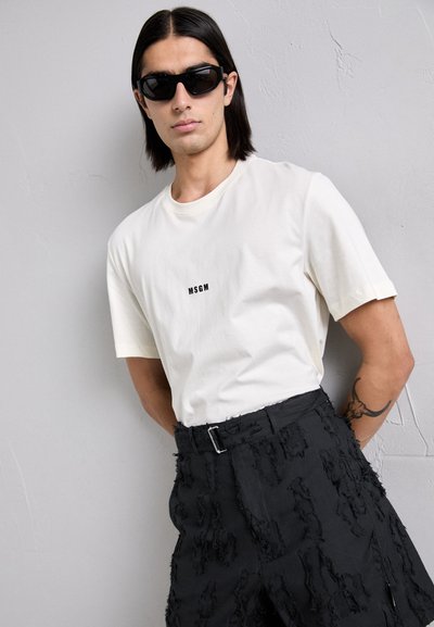 Hvit bomull t-skjorte med svart "MSGM" logo, kombinert med svarte teksturerte bukser med frynser og spenne. Svarte solbriller.