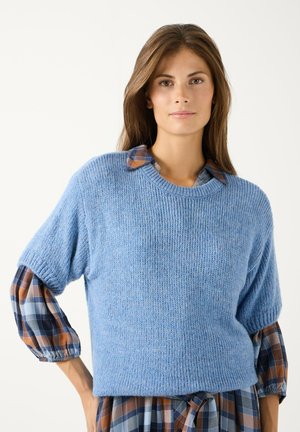 Vrouw met lang bruin haar die een blauwe gebreide trui draagt over een geruite overhemdjurk met blauwe, bruine en witte patronen.