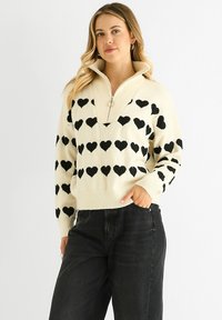 Cream sweater with a quarter-zip collar, featuring black heart patterns, ribbed cuffs, and hem. Paired with high-waisted black jeans.