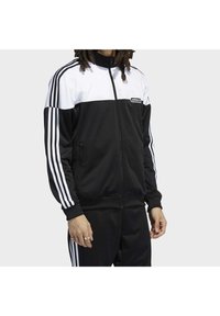 Svart och vit adidas träningsdräkt med dragkedja framtill, med tre vita ränder ner längs varje ärm och på byxans sidor. Slätt tyg.