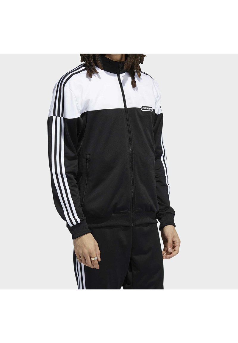 Svart och vit adidas träningsdräkt med dragkedja framtill, med tre vita ränder ner längs varje ärm och på byxans sidor. Slätt tyg.