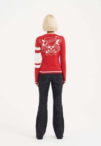 Roter Pullover mit weißen Streifen an den Ärmeln; verfügt über ein Skull-und-Kreuzbones-Design. Kombiniert mit schwarzen Schlaghosen mit strukturierten Mustern.