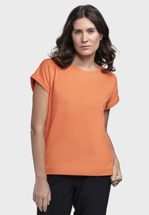 Frau mit welligem dunklem Haar, die ein orangefarbenes Kurzarmshirt und eine schwarze Hose trägt, steht vor einem schlichten hellen Hintergrund.