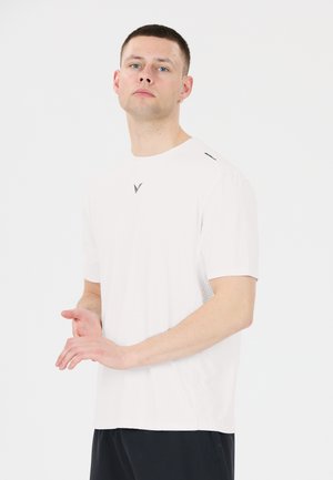 Young man with short hair wearing a white t-shirt and black pants standing against a plain light background.