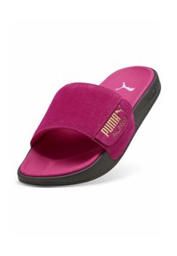 Puma šlepetės su rožiniu zomšiniu dirželiu, tekstūruota rožine pado dalimi ir juoda gumine klase. Dirželyje yra auksinė prekės ženklo žymė.