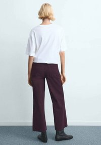 Bela bombažna majica s kratkimi rokavi, kombinirana z visokim pasom, širokimi bordo rdečimi hlačami in črnimi gležnjaki, pogled od zadaj.