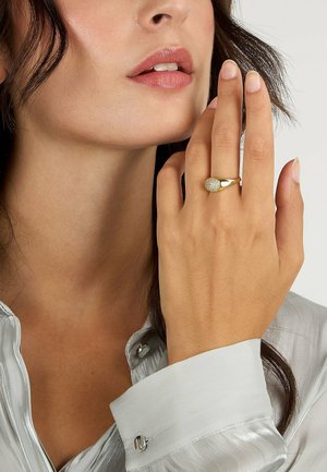 Frau trägt einen goldenen Ring mit kleinen Diamanten am Ringfinger, berührt ihr Gesicht, gekleidet in ein hellgraues Satinkleid.