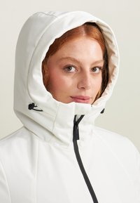 Giacca bianca isolata con un ampio cappuccio, dotata di una cerniera frontale e corde regolabili per una vestibilità sicura. Tessuto liscio e resistente all'acqua.