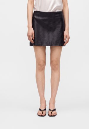 Mujer lleva una minifalda negra texturizada, blusa blanca y sandalias negras con detalle dorado, de pie frente a un fondo claro y sencillo.