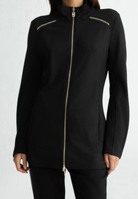 Czarna kurtka na zamek z przodu z wysokim kołnierzem, ozdobiona złotymi elementami, z dwoma kieszeniami na piersi zapinanymi na zamek oraz dopasowanym krojem i długimi rękawami.