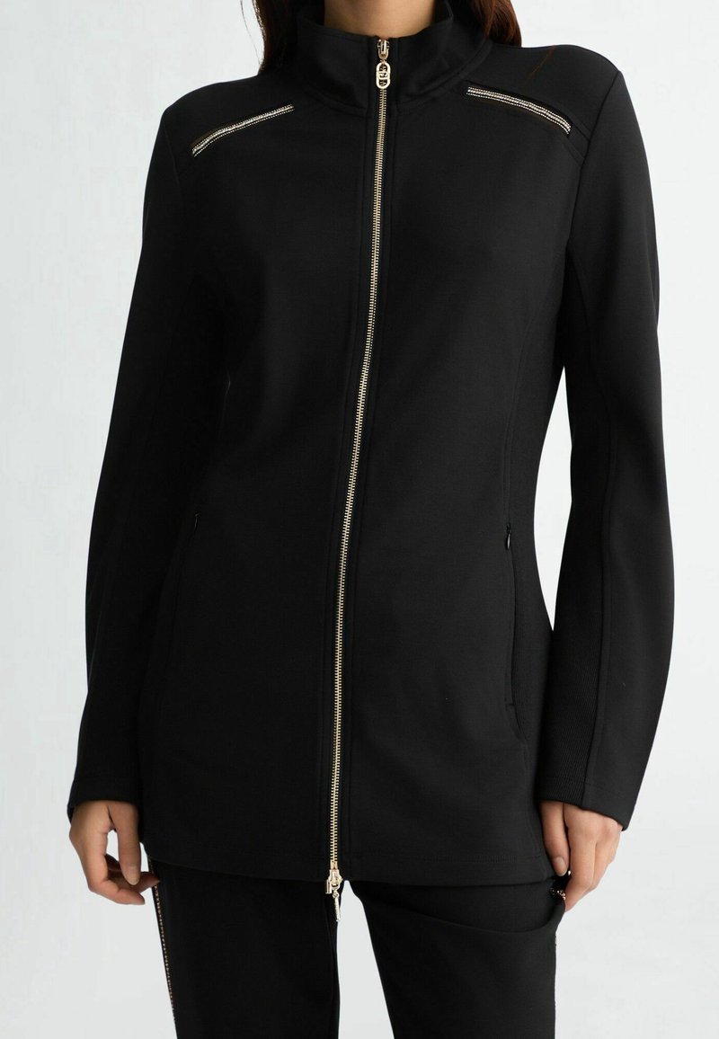 Czarna kurtka na zamek z przodu z wysokim kołnierzem, ozdobiona złotymi elementami, z dwoma kieszeniami na piersi zapinanymi na zamek oraz dopasowanym krojem i długimi rękawami.