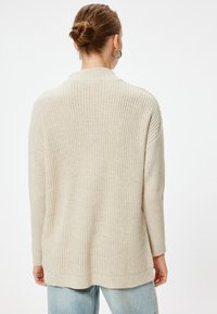 Beiger Strickpullover mit lockerer Passform, ausgestattet mit einem Schalkragen und langen Ärmeln. Die Textur ist gerippt, mit einer leicht übergroßen Silhouette.