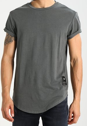 Homme portant un T-shirt gris foncé à col rond avec les manches retroussées et une petite étiquette noire sur le côté inférieur gauche, assorti à un pantalon sombre.