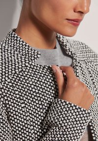 Schwarz-weiße Hahnentritt gemusterte Jacke mit kragenähnlichem Ausschnitt, die strukturierter Stoff zeigt und eine Nahaufnahme eines grauen Pullovers darunter.