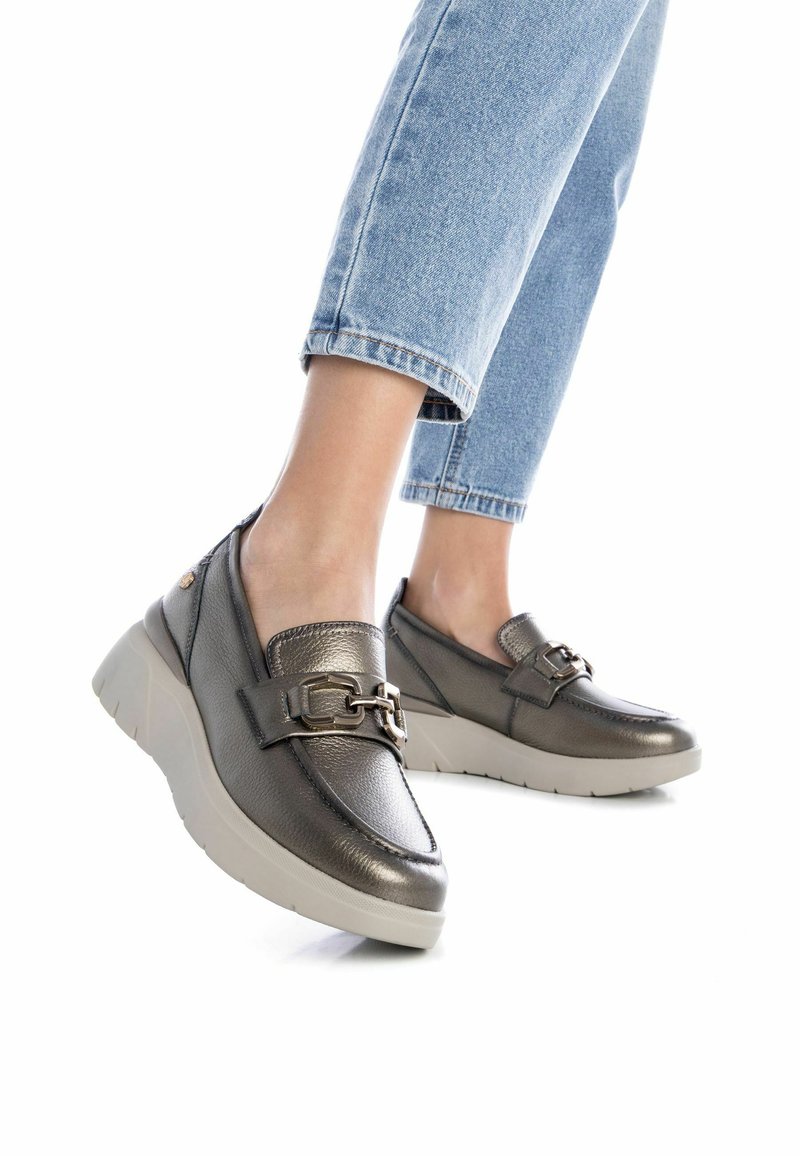 Zapatos slip-on de color gris metálico con una suela blanca gruesa. Cuentan con una hebilla decorativa y una textura suave. Se combinan con jeans de tiro bajo de color azul claro.