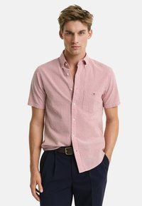 Kurzarm-Hemd für Herren in rot-weißen Streifen, aus Baumwolle, mit Button-Down-Kragen, Brusttasche, getragen mit navyblauen Hosen und braunem Gürtel.