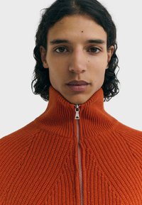 Persona con cabello oscuro rizado que lleva un suéter de punto naranja con cremallera, mirando hacia adelante frente a un fondo claro y liso.