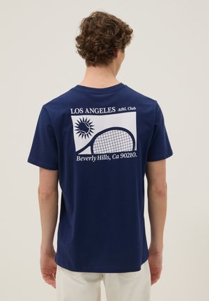 Giovane uomo con capelli ricci che indossa una maglietta blu navy con una grafica bianca di sole e racchetta da tennis con la scritta "Los Angeles Athl. Club".