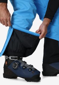 Blauwe waterdichte skibroek met een ritsopening die een zwarte voering onthult. Een zwarte en blauwe skibot is zichtbaar, met verstelbaar materiaal.