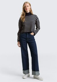 Pull en maille rayé bleu marine et crème avec un col montant, associé à un jean large foncé avec des revers, et des baskets blanches.