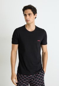 Schwarzes Baumwoll-T-Shirt mit kurzen Ärmeln, runder Halslinie und kleinem roten "HUGO"-Logo auf der Brust, kombiniert mit gemusterten schwarzen Shorts.