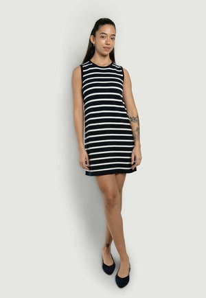 Frau steht und trägt ein ärmelloses schwarz-weiß horizontal gestreiftes Kleid und schwarze Ballerinas, mit einem floralen Tattoo am linken Unterarm und Knöchel.