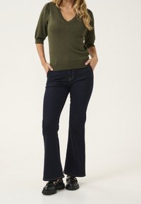 Olivgrön V-ringad tröja med korta puffärmar, kombinerad med mörkblå utsvängda jeanstassar och svarta klumpiga skor.