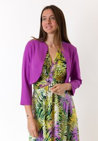 Giacca corta viola sopra un vestito verde e giallo con motivo a palme e vita legata. Le mani del modello mostrano numerosi anelli.