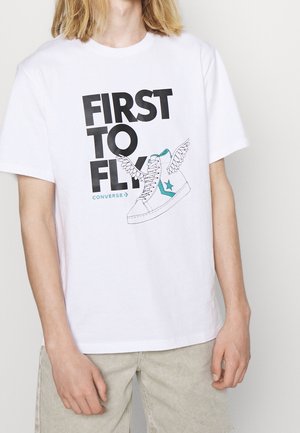 Biała bawełniana koszulka z dużą czarną i tealową grafiką, która zawiera tekst "FIRST TO FLY" oraz sneakersy z skrzydłami.