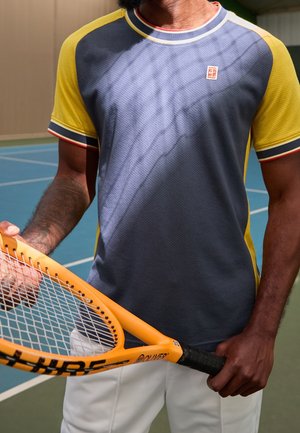 Mand holder orange tennisracket på indendørs bane iført gul og blå kortærmet skjorte og hvide shorts, viser tatoveret arm.