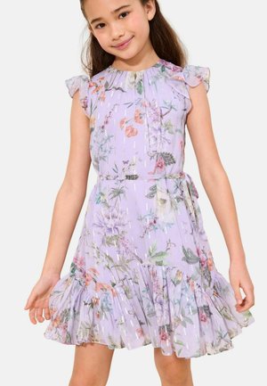 Mädchen trägt ein ärmelloses lavendelfarbenes Blumenkleid mit Rüschensaum und Taillengürtel, lächelt vor schlichtem Hintergrund.