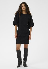Mini vestido negro texturizado con mangas cortas abullonadas, escote redondo y silueta ajustada. Combinado con botas negras de tacón alto hasta la rodilla.
